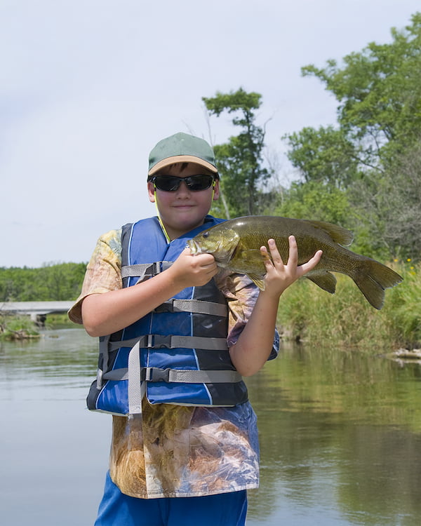 Boy in a life jacket holding a bass fish by a pond, showcasing recreational fishing and healthy aquatic ecosystems.