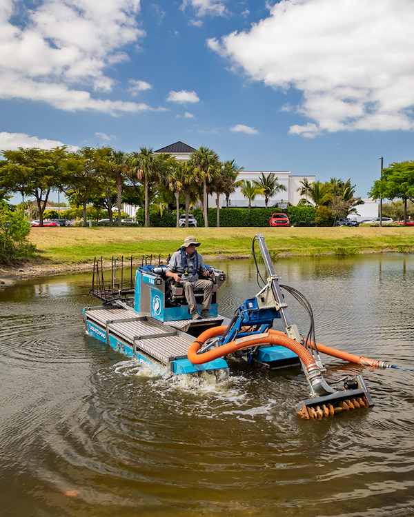 Amphibious hydraulic dredging machine, Truxor T-50, operating in pond, removing sediment and organic muck, with operator seated, surrounded by trees and clear blue sky, emphasizing low-impact dredging for water quality improvement.